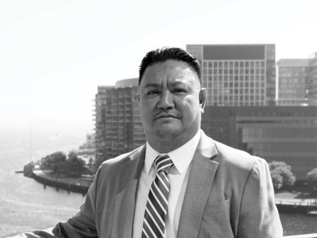 A man in a suit stands on a balcony with a cityscape and waterfront in the background, captured in black and white.