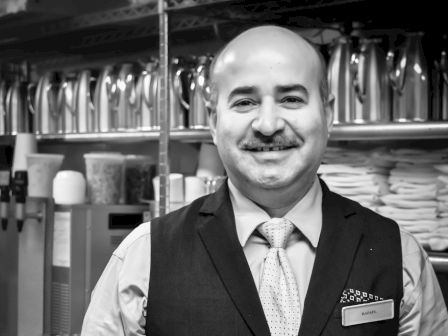 A man is standing and smiling in what appears to be a professional or service environment, dressed in a vest and tie, likely a server or bartender.