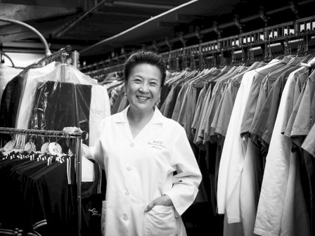 A person in a lab coat standing and smiling, surrounded by rows of hanging garments in what appears to be a dry cleaning facility.