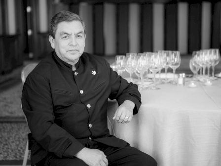 A person dressed in formal attire sits at a table with numerous wine glasses arranged neatly.