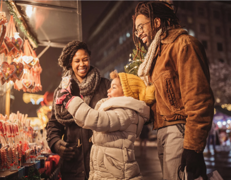 Three smiling adults and a baby at a festive market stall, admiring heart-shaped decorations under twinkling lights, joyful family moment.