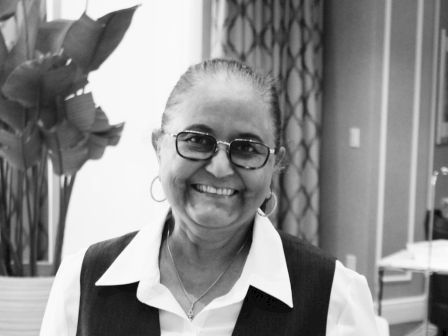 A smiling person wearing glasses and a uniform with a name tag stands indoors, with plants and decorative elements in the background.