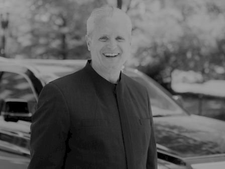 A smiling man in a dark suit stands in front of a vehicle with trees blurred in the background. The image is in black and white.
