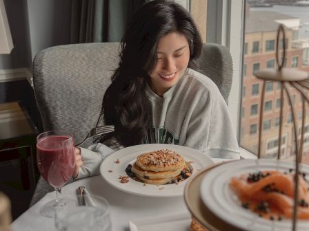 A woman sits at a restaurant table with pancakes, coffee, and a glass of pink drink, looking down at her plate near a window.