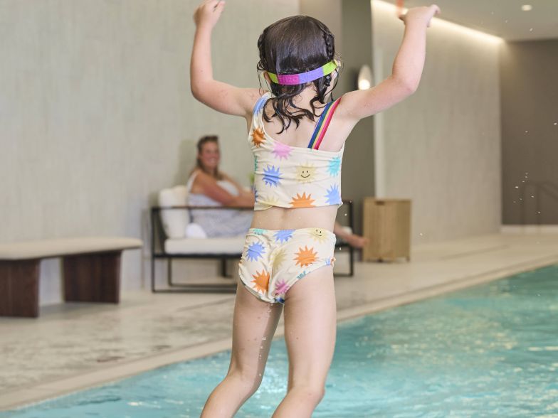 A child wearing a floral swimsuit leaps off a pool edge, mid-air, inside an indoor swimming area, with a person watching from a bench in the background.
