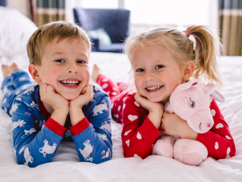 Two smiling children in matching pajamas lie on a bed with their heads resting on their hands, looking at the camera.
