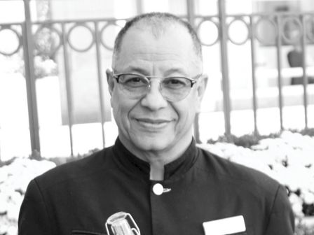 A man in a uniform is holding a bottle of water with a smile, standing outdoors with a fence and plants in the background.