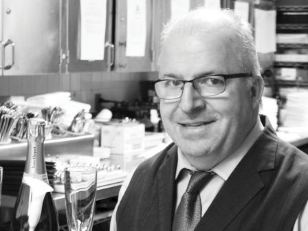 A suited man with glasses, holding a tray with a wine bottle and two glasses, stands in a kitchen-like setting, smiling.