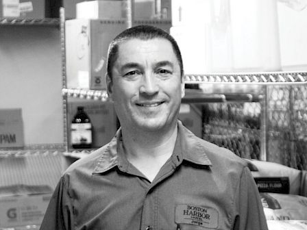 A man in a uniform, smiling, stands in a room filled with boxed and packaged items on shelves.