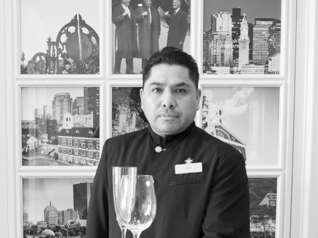 A uniformed person holds a tray with two glasses in front of a wall displaying various framed black-and-white photographs, some showing landmarks.