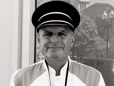 A black and white photo of a man wearing a uniform and a hat, standing and smiling, with a building in the background.
