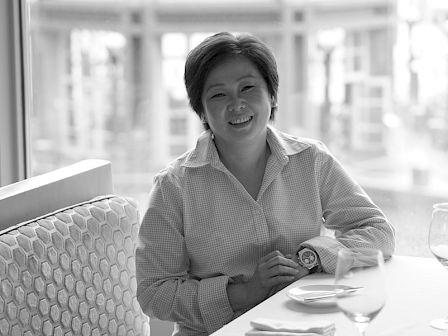 A person smiling and sitting at a table in a restaurant with a building visible through the window in the background.