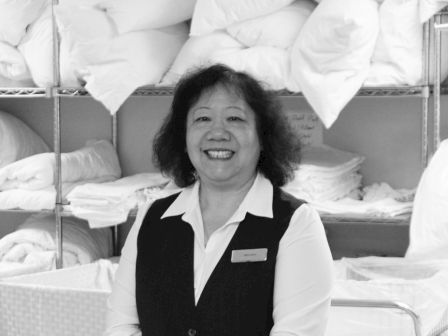A smiling person in uniform stands in front of linen shelves loaded with folded towels and bedding in a housekeeping or laundry setting.