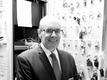 A man in a suit stands smiling in front of a wall with many keys hanging, likely indicating a security or management role.