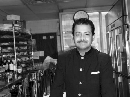A person in a uniform stands in a kitchen, smiling at the camera. The background features shelves and a catering setup with dishes and cutlery.