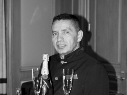 A waiter holding a tray with a bottle of champagne and two champagne glasses. The background features a formal room setting.