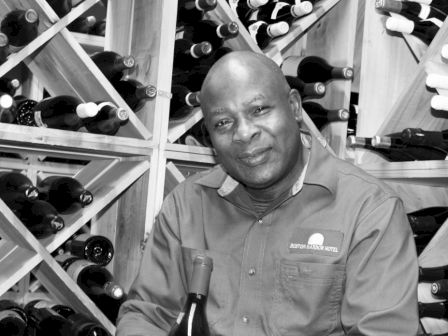 A person is seated in a wine cellar, holding a bottle of wine, with shelves of wine bottles in the background.