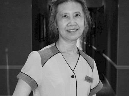A person is standing in a hallway. They are smiling and wearing attire that might be work-related. The photo is in black and white.