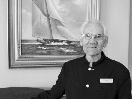An older man in a black uniform stands in front of a painted sailboat picture, with one hand on a covered table, smiling at the camera.