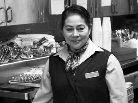 A person is standing in a kitchen or work area with various utensils and items on the counter, wearing a uniform and smiling at the camera.