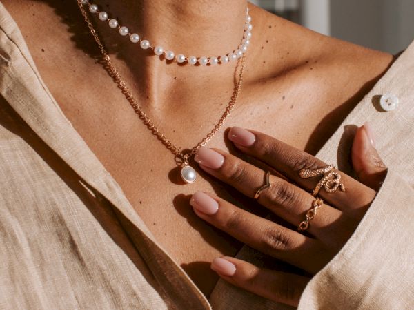 A person wearing a beige shirt, pearl earrings, a pearl choker, and a pendant necklace. Their hand adorned with rings and light pink nail polish.