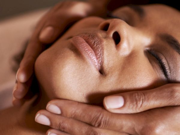 A person receives a gentle facial massage, hands placed on their face and neck, eyes closed in relaxation.