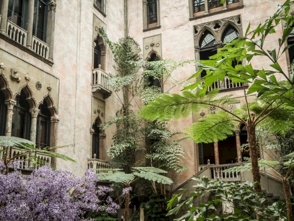 The image shows an old building with arched windows surrounded by lush greenery and purple flowers, creating a serene and historic atmosphere.