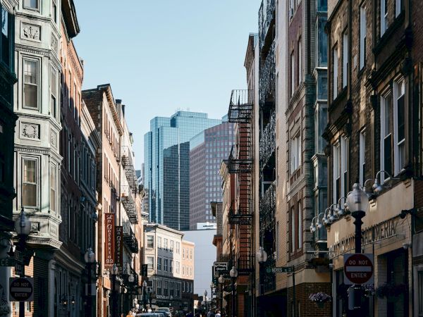 The image depicts an urban street with historic buildings, narrow pathways, and a bakery sign on the right. Modern skyscrapers are visible in the background.
