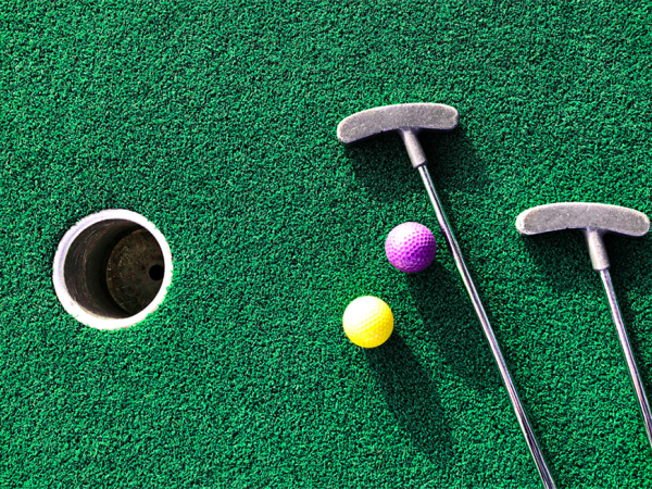 The image shows a mini-golf hole on green turf, with two putters and two golf balls, one yellow and one purple, placed near the hole.