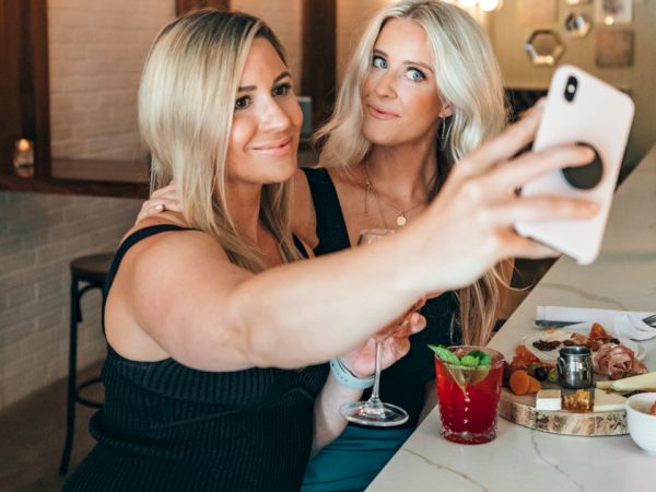 Two women are taking a selfie at a bar. One holds a drink, and there are various snacks and drinks on the table.