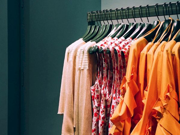 A row of colorful clothes, including white, patterned, and orange outfits, neatly hang on a metal clothing rack in a retail store.