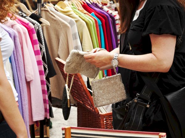 Two people are at an outdoor clothing market; one holds a beige clutch, while various colorful garments hang on a rack in the background.