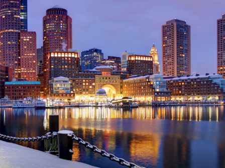 A snowy waterfront skyline at dusk, with tall buildings glowing, harbor lights reflecting on calm water, and a chain-roped promenade in foreground.