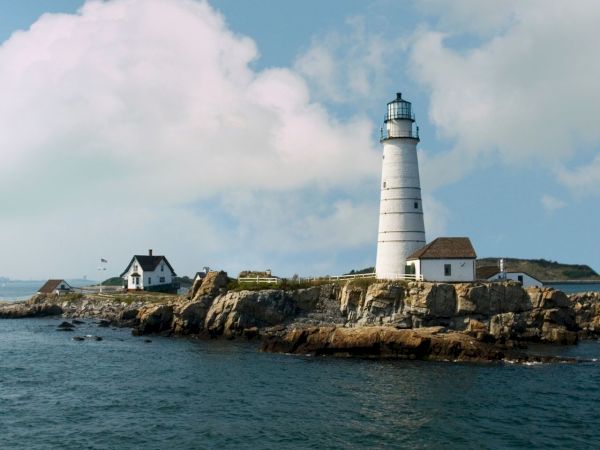 A lighthouse on a rocky island with adjacent small buildings and a cloudy sky in the background sits surrounded by water.