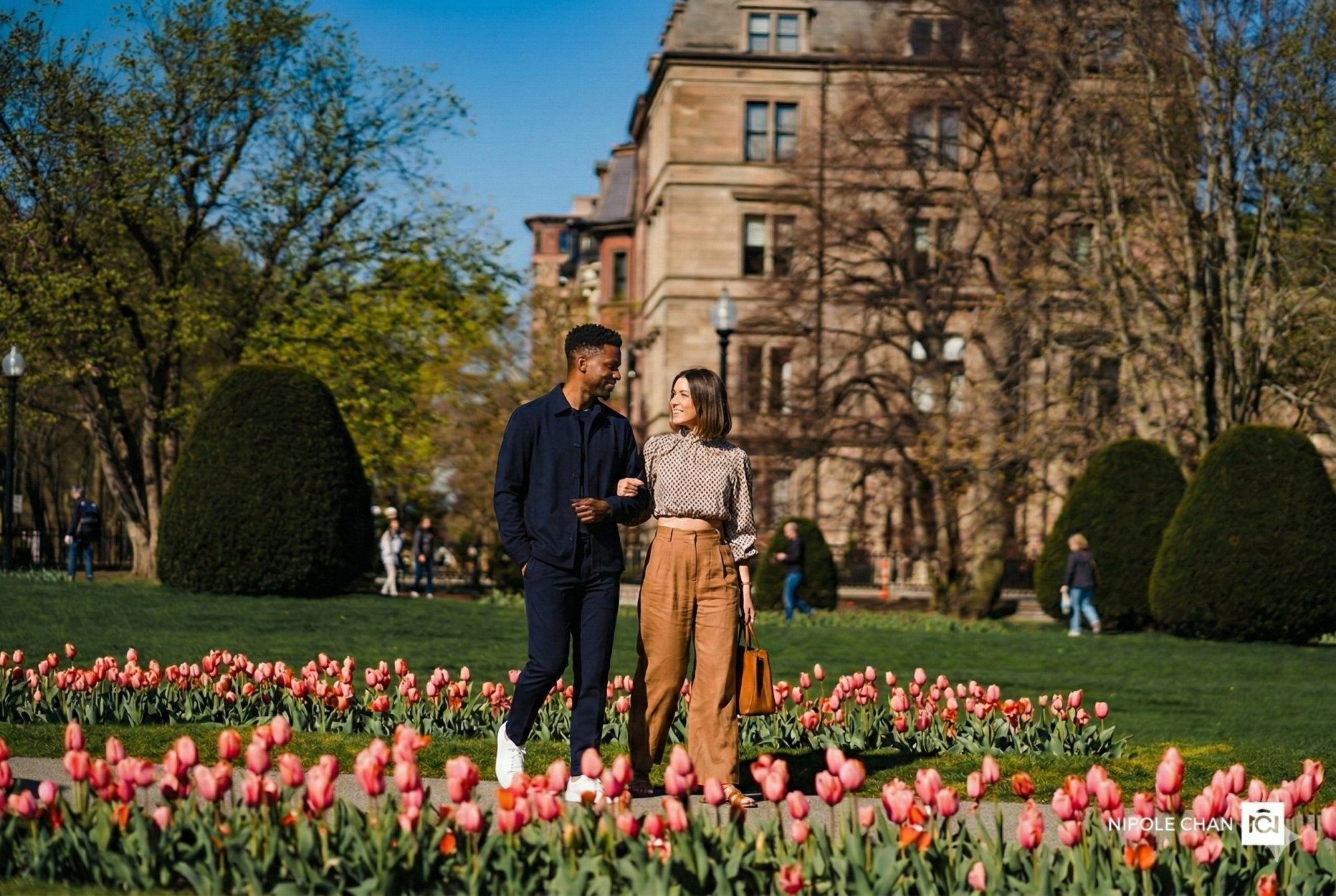 A couple strolls through a tulip garden by a historic brick building, enjoying a sunny day amid green trees and colorful blossoms.