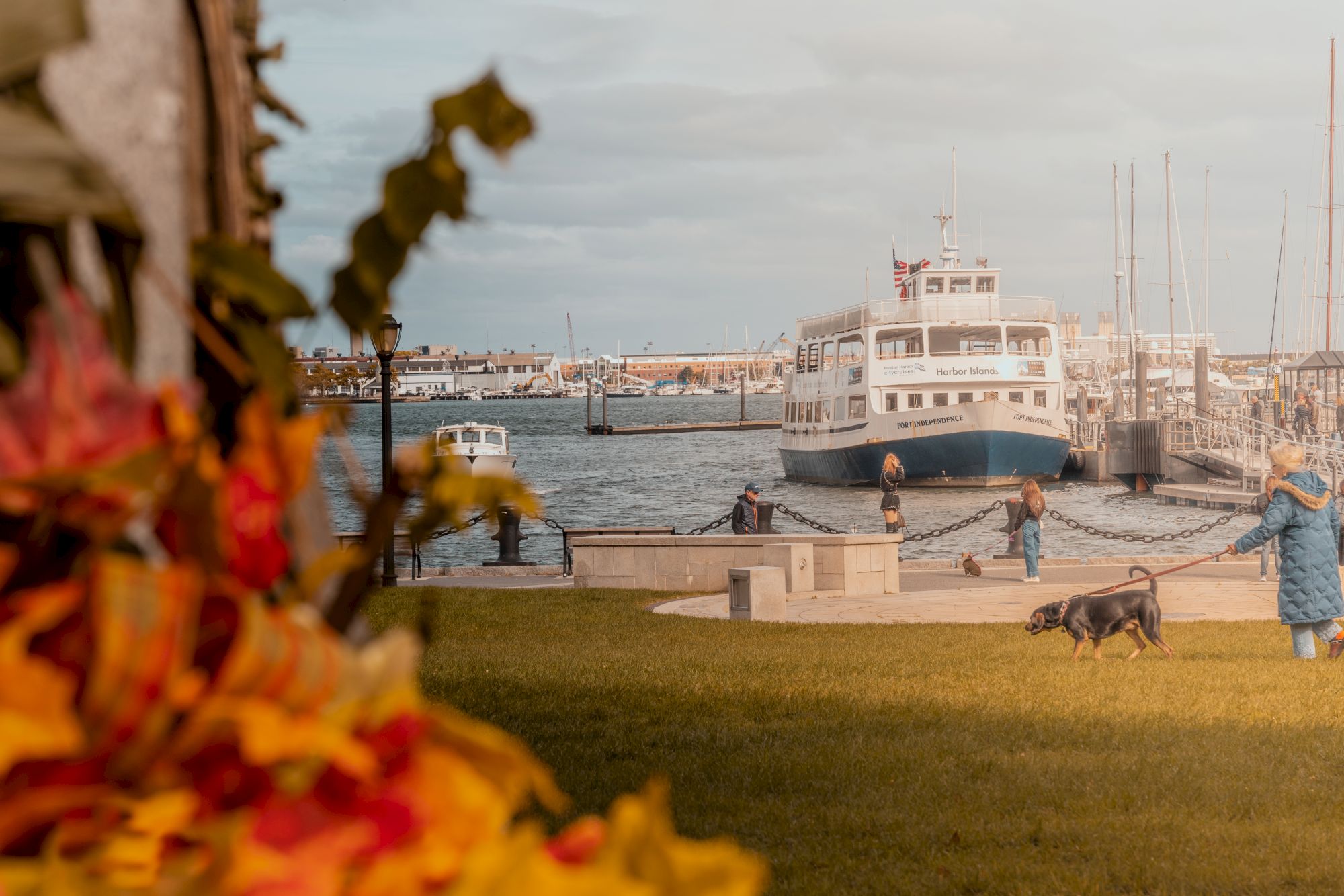 A harbor marina with the ship &ldquo;Fort Independence,&rdquo; sails and docked boats, and people walking dogs along the waterfront.