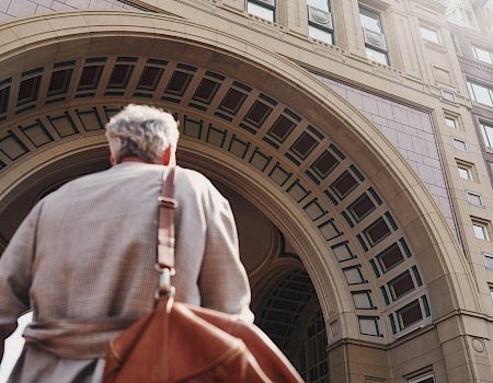 A person with gray hair and a brown bag is walking towards an arched architectural structure made of stone, visible from a low angle.