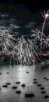 A nighttime scene with bright fireworks bursting over a harbor, boats dotting the water, and a distant city skyline shimmering in the background.