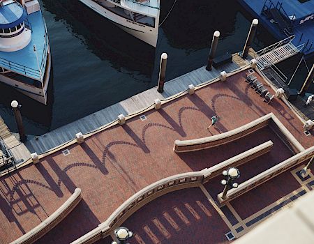 An aerial view of a waterfront promenade with boats docked at the pier, light poles casting shadows, a lone walker, and decorative brickwork.