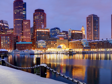 Boston skyline at dusk along the harbor, tall glassy towers glow, reflection on calm water, snow on the foreground, a chain railing guides the scene.
