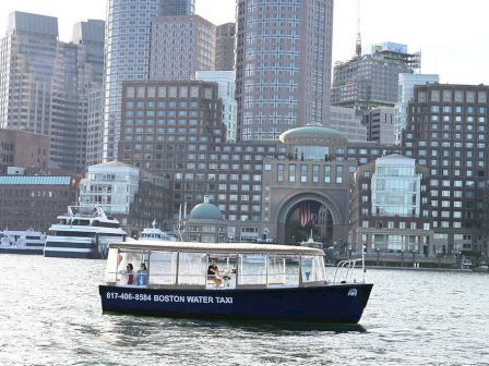 A water taxi labeled "Boston Water Taxi" is moving on water with a cityscape in the background, featuring tall buildings.