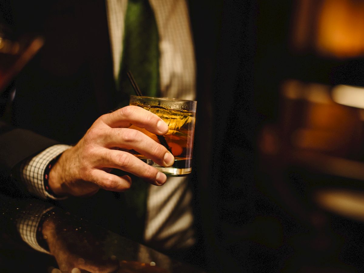 A person in a suit holds a glass of whiskey or whiskey on the rocks at a dim bar, toasting or relaxing in a low-lit setting.