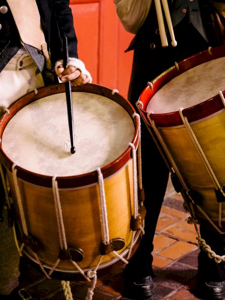 Two individuals are holding and playing traditional drums, dressed in historical attire.