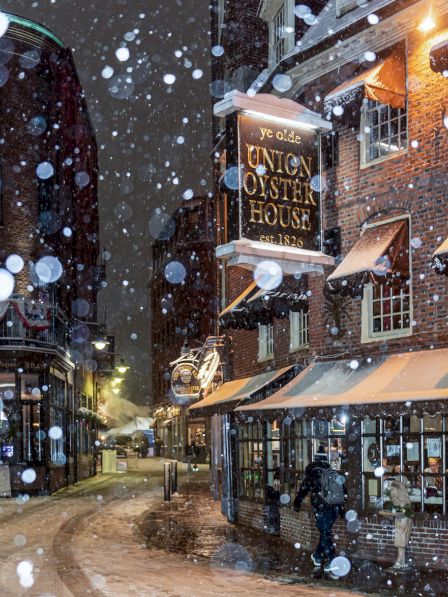 Snowy street scene with old-town buildings, a pub sign &ldquo;Union Oyster House,&rdquo; snow falling, warm lights glow from windows, cozy atmosphere.