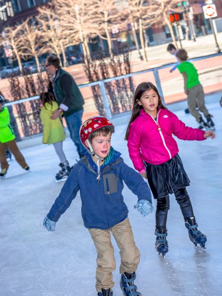 Two kids skate with others gliding on an outdoor rink; a girl in pink leads while a boy in blue follows, all in winter attire.