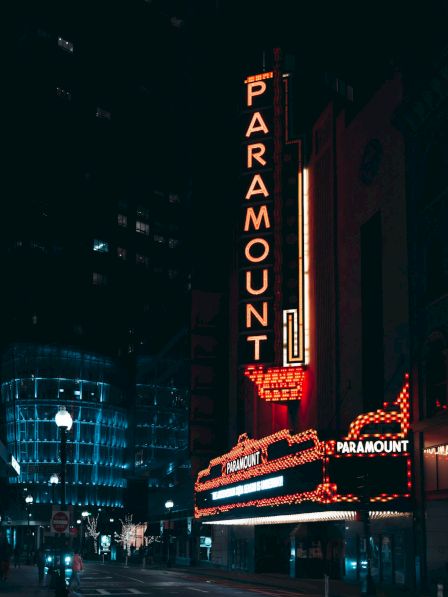 Neon-lit theater sign reads "PARAMOUNT" in a city street at night, surrounded by illuminated buildings and a dark sky.