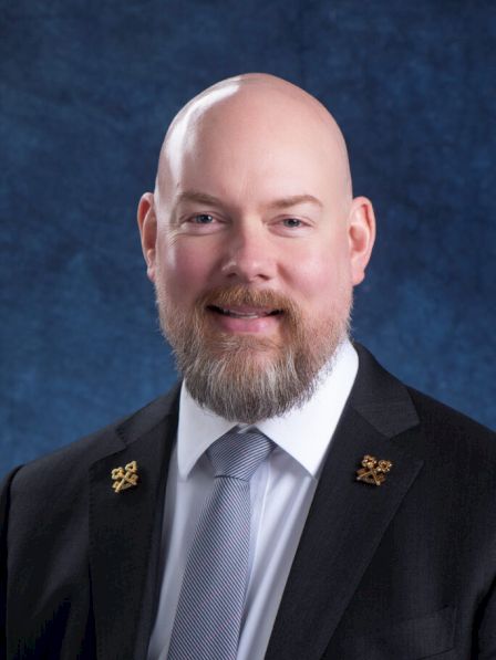 A man with a beard is wearing a suit, tie, and pins on his lapels, standing against a solid blue background.