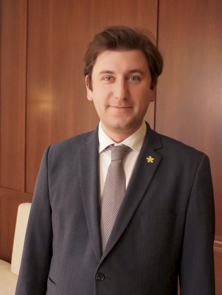 A person in a suit and tie stands in front of a wooden wall with partially visible letters.