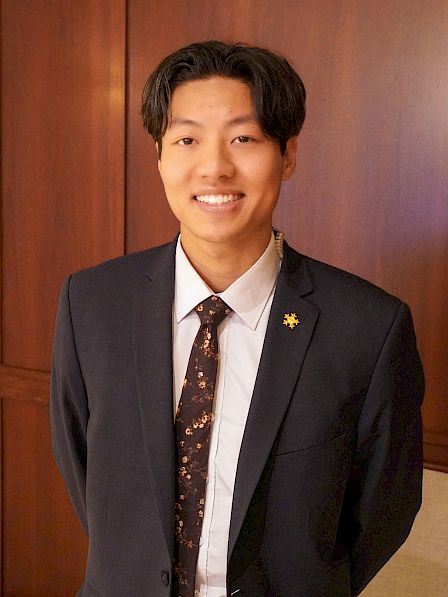 A person in a suit stands indoors, smiling with hands behind their back against a wooden panel background.
