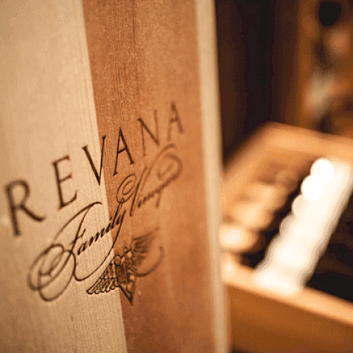 The image shows a close-up of a wooden surface with the "Revana Family Vineyard" logo and a blurred background of a wine display or collection.